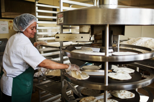 A woman in a hairnet and gloves operates a machine making tortillas in a commercial kitchen.