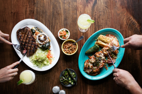 A table with two plates of Mexican food, drinks, and hands holding utensils, ready to eat.