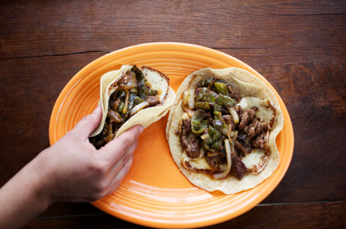 A hand holds a tortilla filled with beef and peppers, next to another similar tortilla on an orange plate.