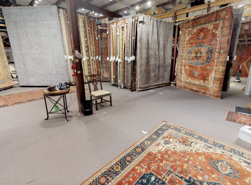 A spacious rug showroom featuring various large rugs displayed on walls and the floor, with a chair and table in the foreground.