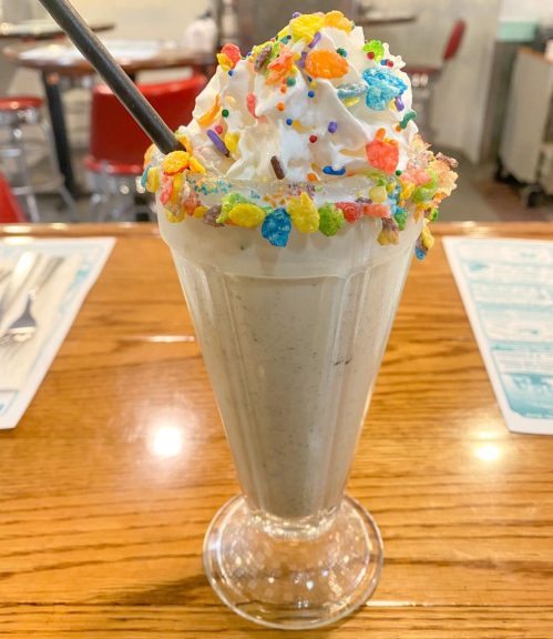 A tall milkshake topped with whipped cream and colorful cereal sprinkles, served in a classic glass.