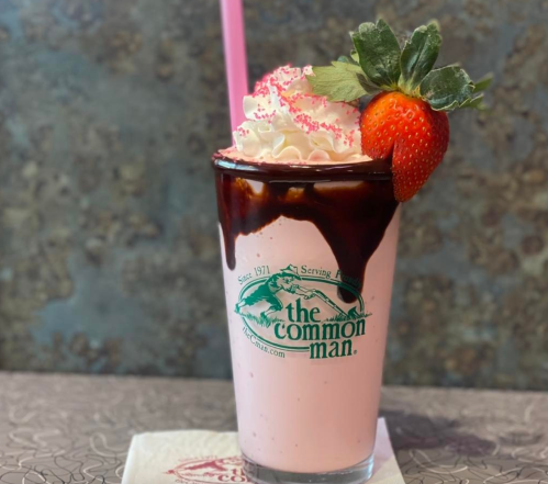 A pink milkshake topped with whipped cream, chocolate drizzle, a strawberry, and pink sprinkles in a branded glass.