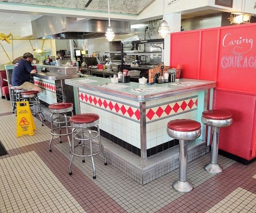 A retro diner interior featuring a counter with stools, a kitchen area, and a caution sign on the floor.