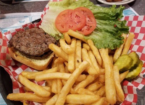 A burger with a beef patty, lettuce, and tomato on a bun, served with a side of golden French fries and pickles.