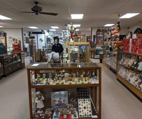 A vintage toy museum filled with glass display cases, dolls, and collectibles, featuring a mannequin in a black outfit.