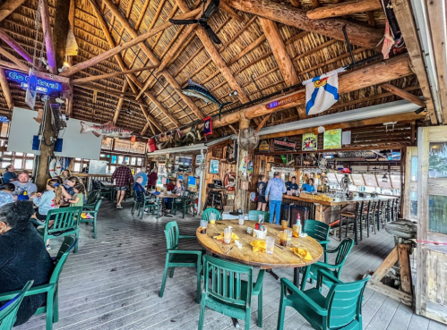 A rustic restaurant interior with wooden beams, thatched roof, and patrons dining at tables. Bar area in the background.