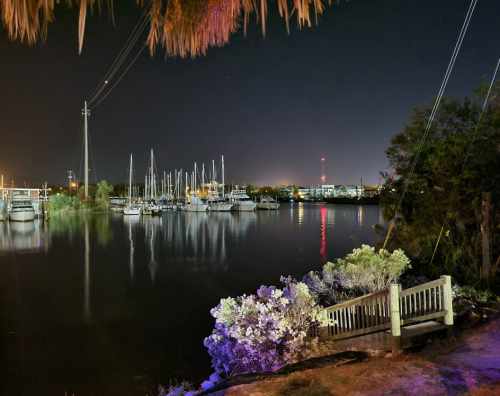 Nighttime view of a calm marina with boats, reflections on water, and colorful lights in the background.