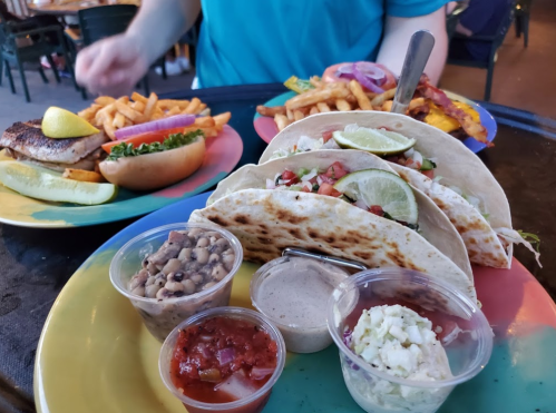 A colorful plate with tacos topped with lime and tomatoes, accompanied by dips, and a background of fries and a sandwich.