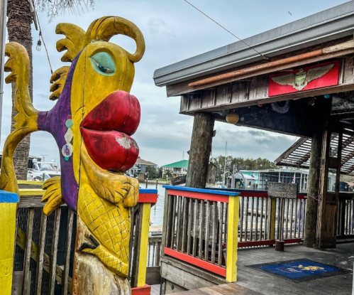 Colorful wooden fish sculpture with exaggerated features near a waterfront building. Overcast sky in the background.