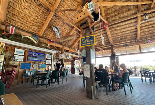 A lively beachside restaurant with a thatched roof, wooden decor, and patrons enjoying drinks at tables.