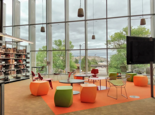 Modern library interior with colorful furniture, large windows, and a view of greenery outside.
