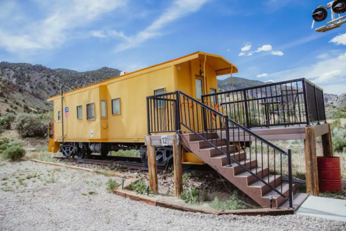 A bright yellow train caboose on a track, featuring a staircase and surrounded by mountains and greenery.