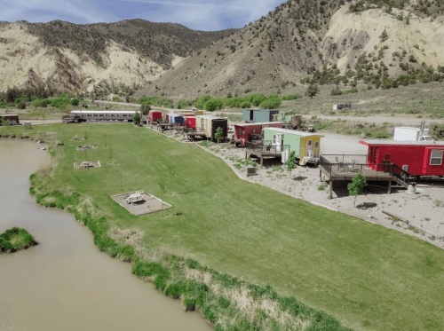 Aerial view of colorful trailers along a river, surrounded by green grass and mountains in the background.