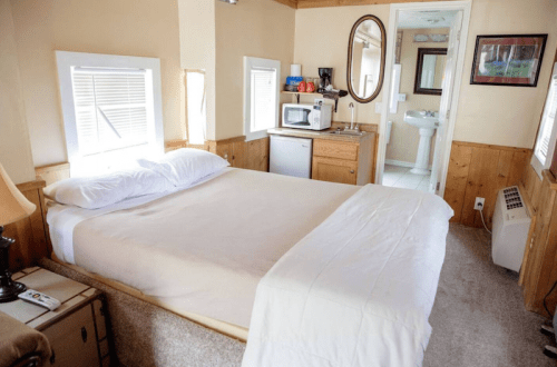 Cozy bedroom with a queen bed, kitchenette, and bathroom visible in the background. Warm, inviting decor.