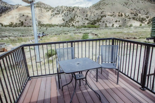A small outdoor deck with a table and two chairs, overlooking a scenic landscape of hills and greenery.