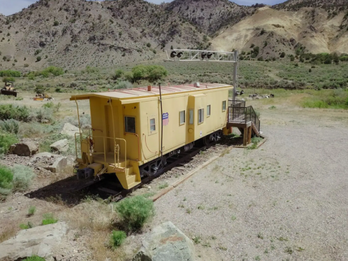 A yellow train caboose on tracks in a rural area, surrounded by mountains and sparse vegetation.