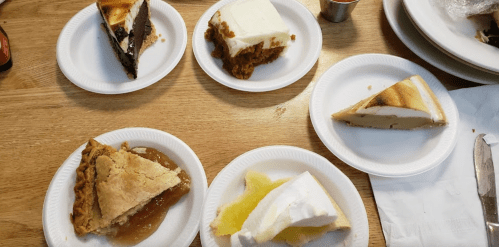 A variety of desserts on paper plates, including chocolate cake, pumpkin pie, cheesecake, and apple pie.