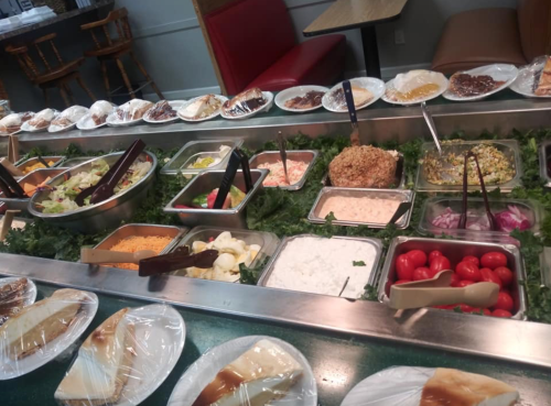 A buffet spread featuring various salads, sides, and desserts in clear containers, with utensils for serving.