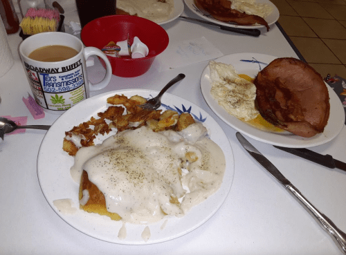 A plate of biscuits and gravy with hash browns, alongside a plate of ham and eggs, with a coffee cup in the background.