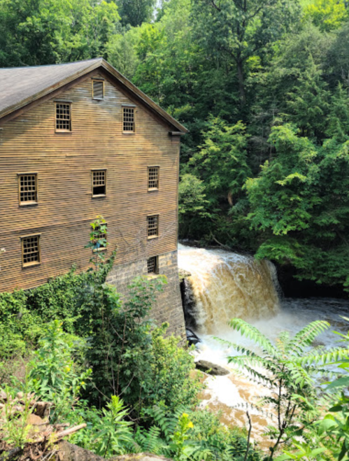 A historic wooden mill beside a cascading waterfall, surrounded by lush green trees and foliage.