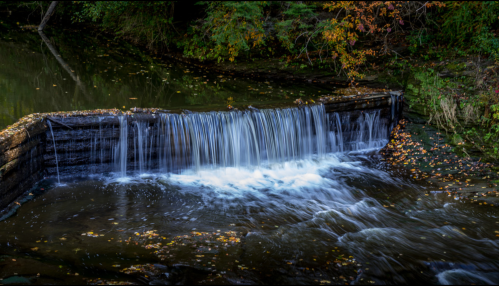 A serene waterfall cascades over a stone ledge, surrounded by autumn foliage and calm waters.