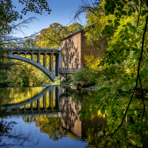 A serene scene featuring a bridge over a calm river, with colorful autumn foliage and a wooden building reflected in the water.