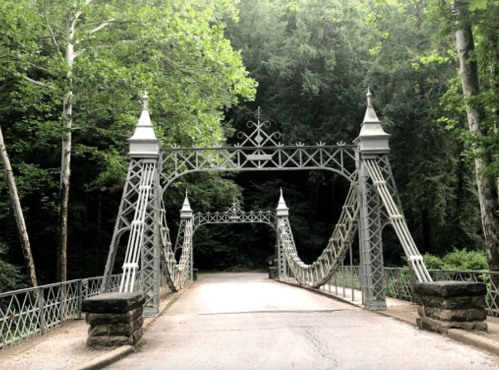 A decorative metal bridge with tall spires, surrounded by lush green trees and a gravel path.