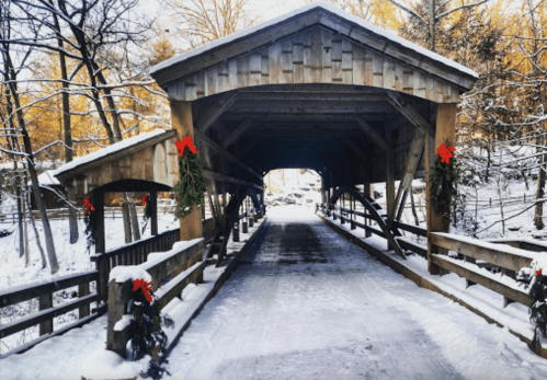 A snow-covered covered bridge adorned with red bows, surrounded by trees in a winter landscape.