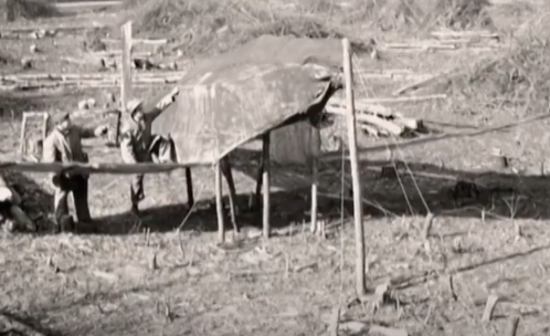 Two people work on a makeshift shelter in a barren landscape, surrounded by logs and debris.