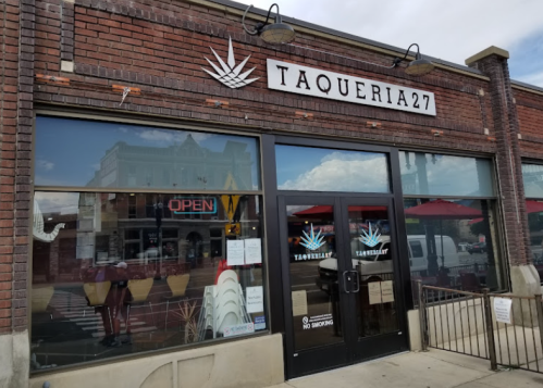 Exterior of Taqueria 27, featuring a brick facade, large windows, and an "Open" sign. Outdoor seating visible.