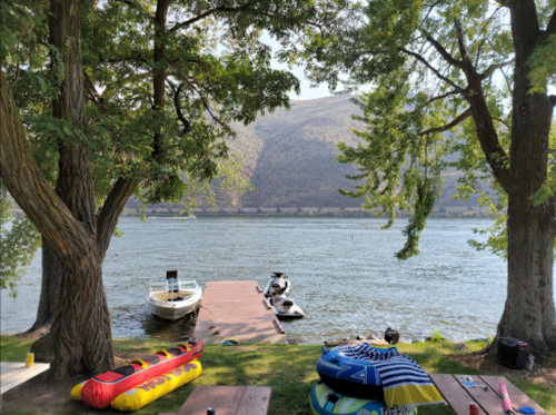 A serene lakeside view with trees, a dock, a boat, and colorful inflatable rafts on the grass.