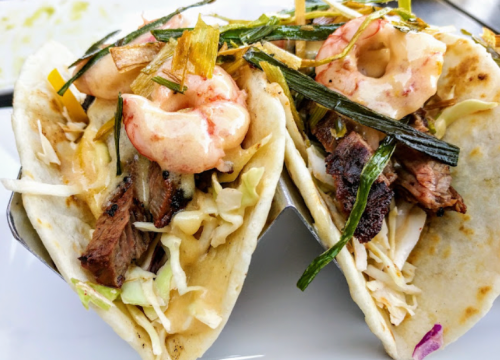 Two tacos filled with shrimp, grilled onions, and cabbage, served in a taco holder.