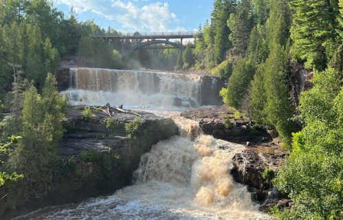 A scenic view of a waterfall cascading over rocks, surrounded by lush greenery and a bridge in the background.