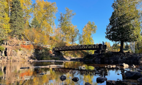 A wooden bridge spans a calm river, surrounded by autumn trees and clear blue skies. Reflections shimmer on the water.