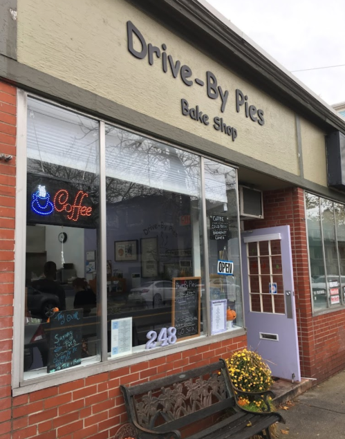 A cozy bake shop named "Drive-By Pies" with a coffee sign, open door, and a bench outside.