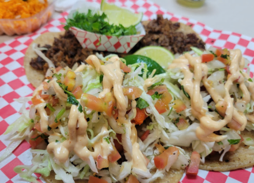Two tacos topped with lettuce, tomatoes, and sauce, served with lime and a side of seasoned meat on a checkered plate.