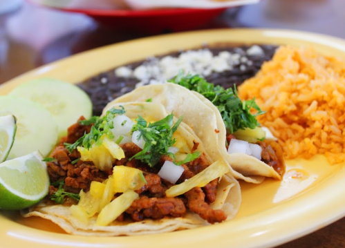 Two tacos filled with meat, pineapple, and cilantro, served with rice, black beans, and lime wedges on a yellow plate.