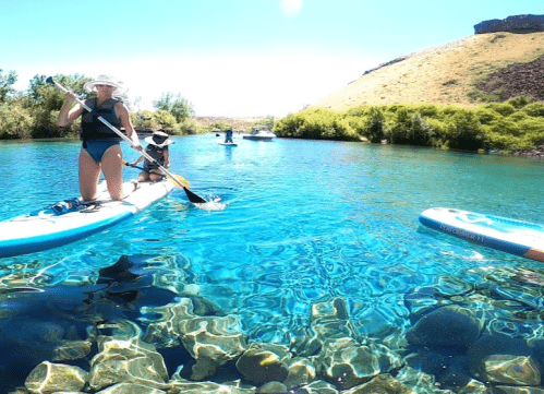 Two people paddleboarding on clear blue water with rocky bottom, surrounded by greenery and a hillside in the background.