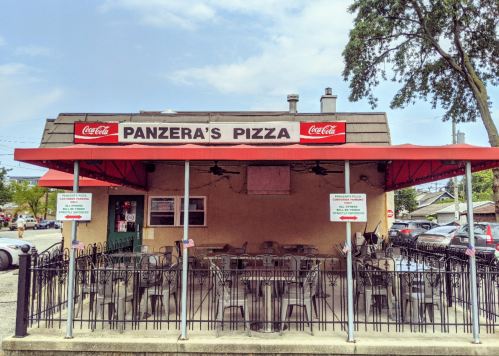 Exterior of Panzera's Pizza with outdoor seating, red awning, and Coca-Cola signage on a sunny day.