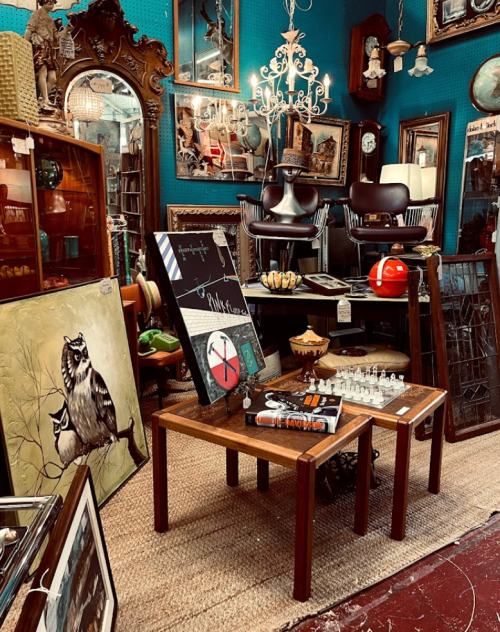 A cozy antique shop filled with vintage decor, mirrors, furniture, and artwork against a teal wall.