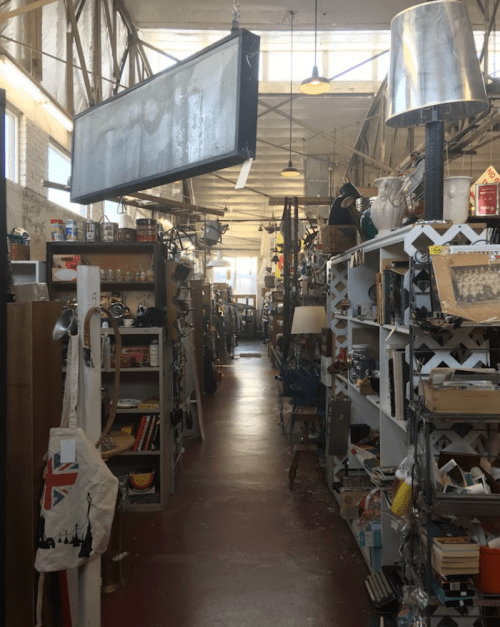 A narrow aisle in a vintage shop filled with shelves of eclectic items and a large hanging sign above.