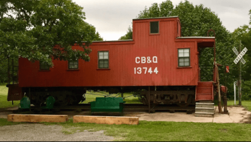 A red caboose labeled "CB&Q 13744" parked on a grassy area, surrounded by trees and a railroad crossing sign.