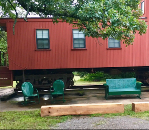 A red train car on wheels with green chairs and a bench nearby, set in a grassy area under a tree.