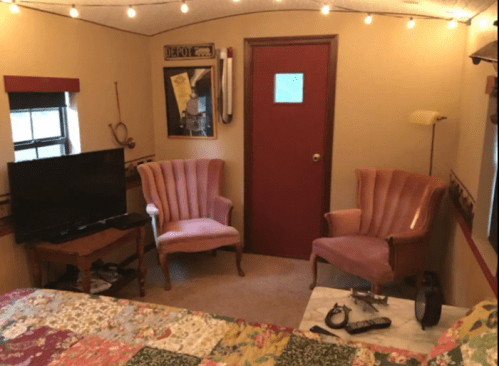 Cozy room with two pink armchairs, a TV, and a bed, decorated with string lights and a red door.