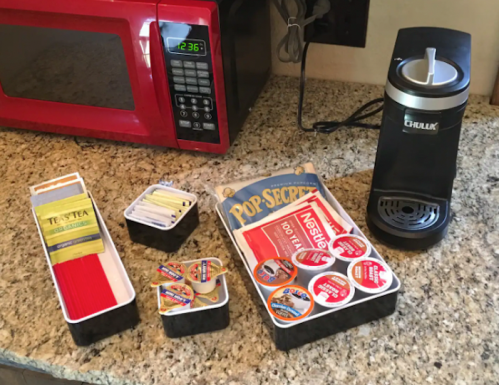 A countertop with a microwave, coffee maker, and containers of tea, popcorn, and sweeteners.