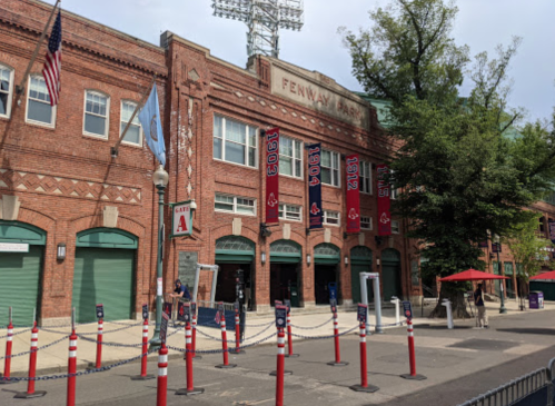 Exterior view of Fenway Park with banners, flags, and a row of red and white barriers in front.