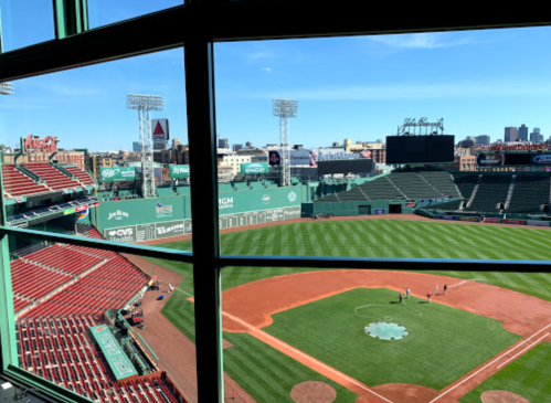 View of Fenway Park from a window, showcasing the green field, empty red seats, and a clear blue sky.