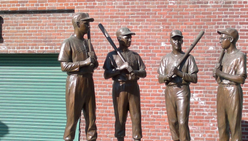 Four bronze statues of baseball players holding bats, set against a brick wall.