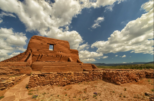 Ancient adobe ruins under a blue sky with fluffy clouds, surrounded by a stone wall and desert landscape.