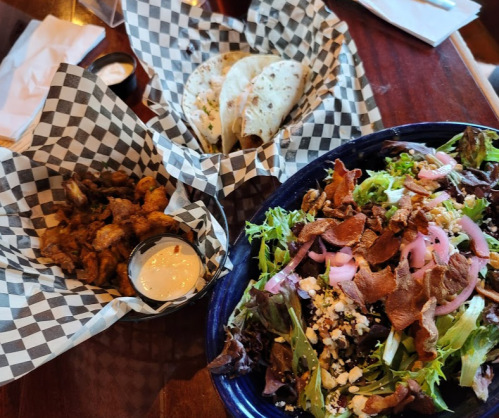 A bowl of salad with greens, bacon, and pickled onions, alongside fried chicken and tortillas in checkered paper.